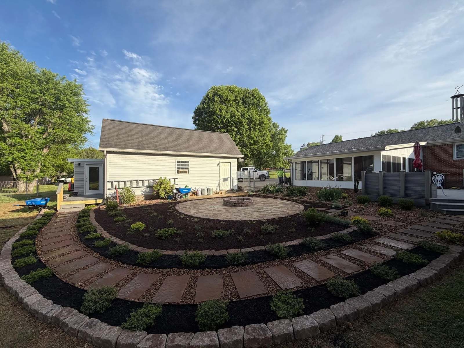 Backyard with circular brick patio, raised garden beds, and a small white shed under sunny skies