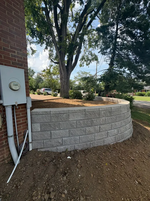 Backyard retaining wall beside a brick house, with a large tree and dirt landscaping.