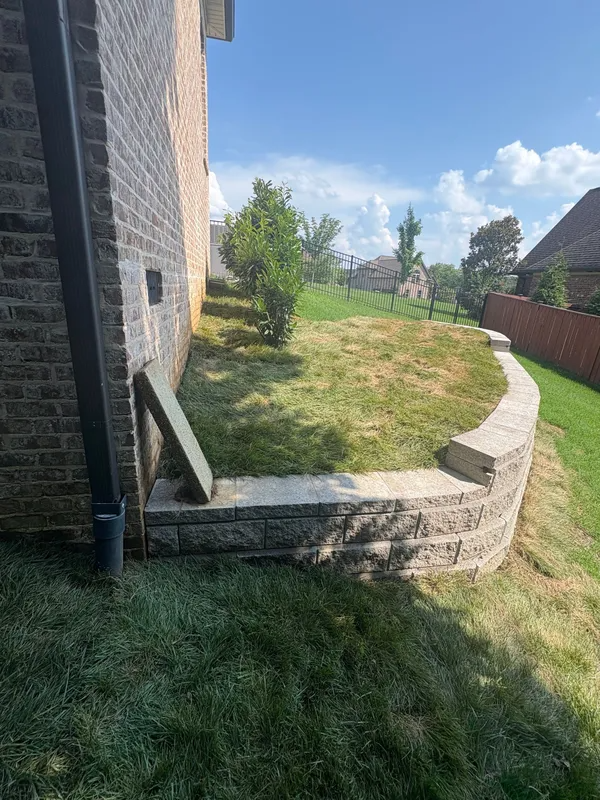 Side yard with a stone retaining wall, grassy slope, brick house, and blue sky on a sunny day