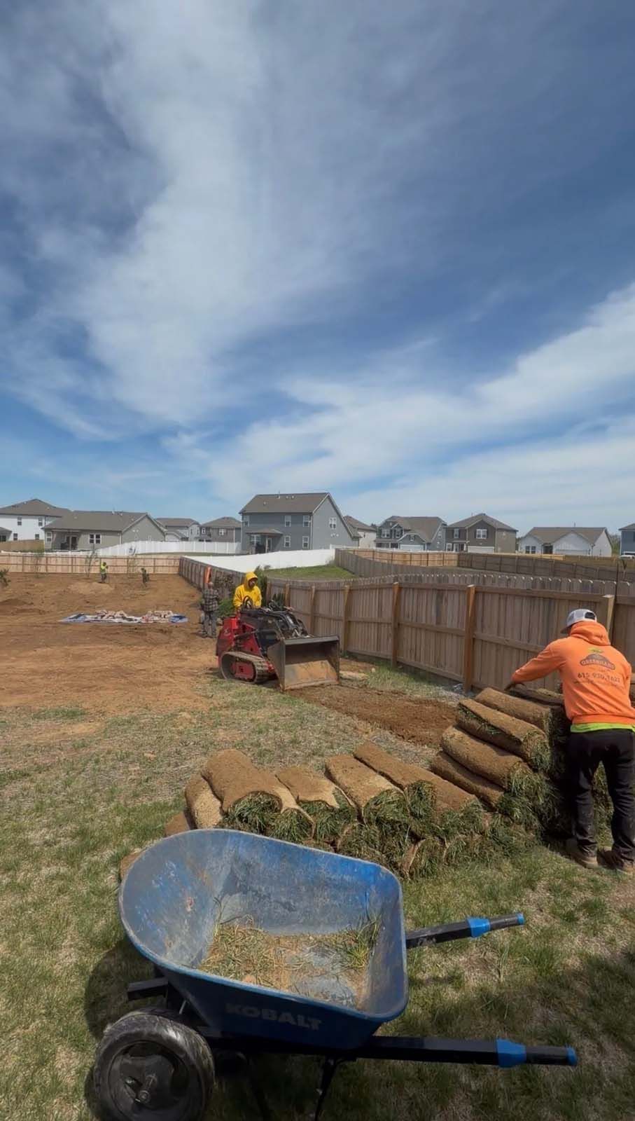 Workers installing a wooden fence in a suburban backyard with a wheelbarrow in the foreground