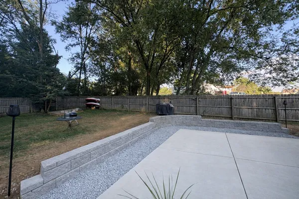 Backyard with concrete patio, gravel retaining wall, grass, trees, and a wooden fence at dusk