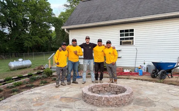 Five people in yellow shirts stand by a circular stone fire pit in a backyard beside a white house.
