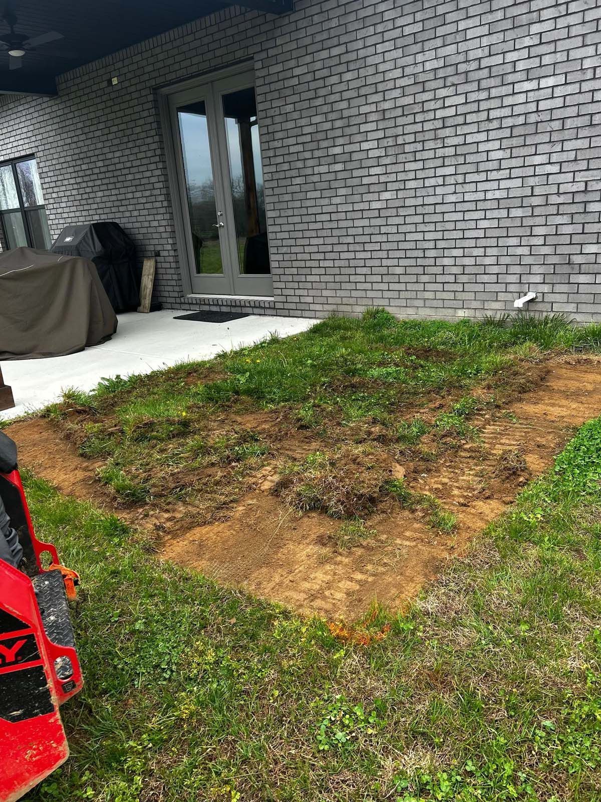 Patchy grass lawn beside a gray brick house with patio doors and a concrete patio.