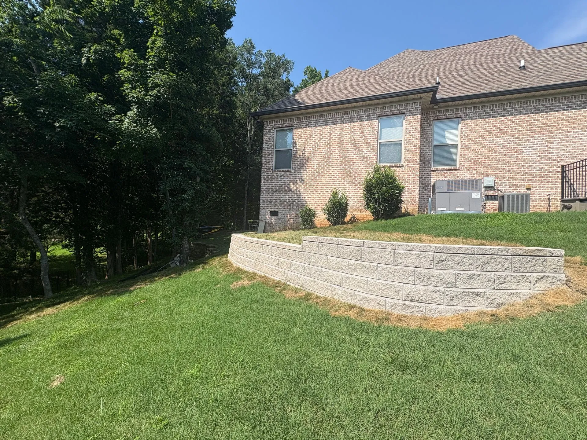 Brick house with stone retaining wall and green lawn beside trees in sunlight