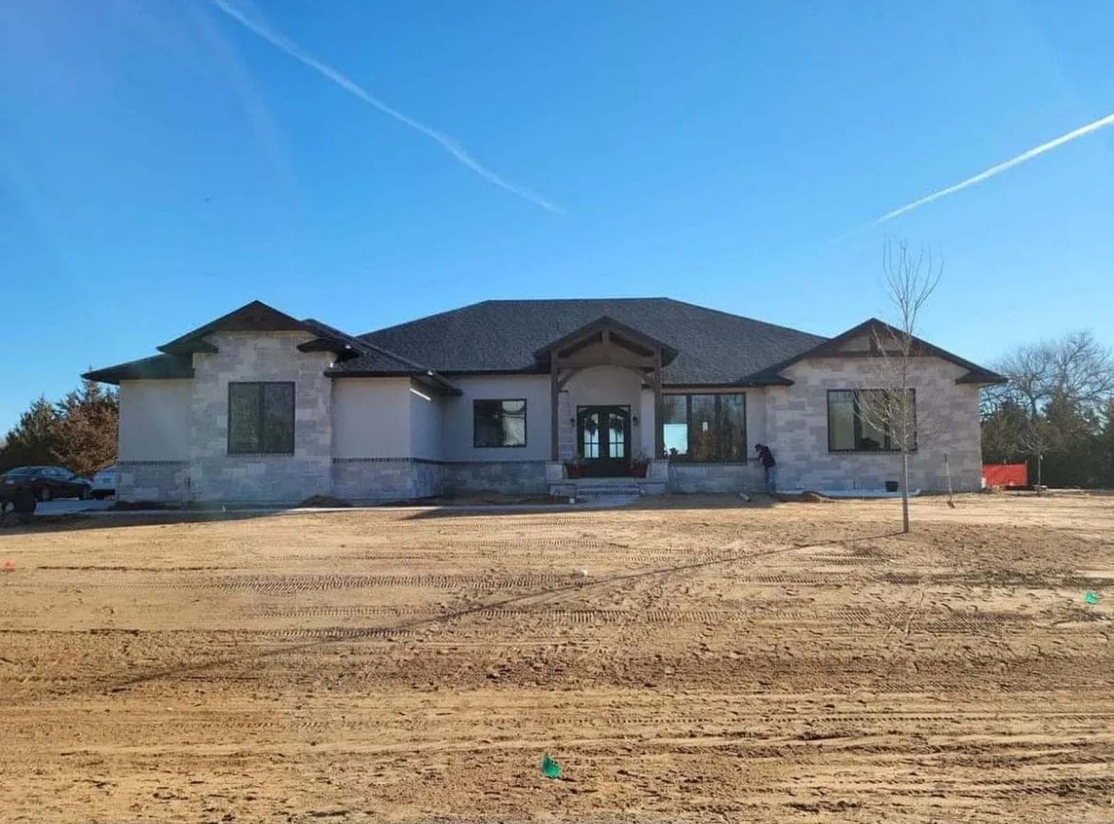 Single-story house under construction with a gray roof and bare dirt front yard under a blue sky