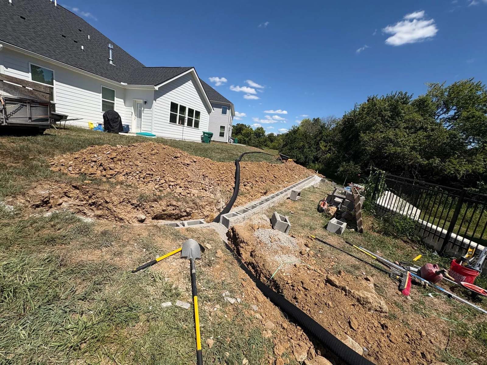 Backyard excavation beside a white house, with exposed trench and a sloped dirt retaining area