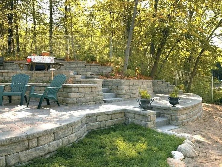 Stone patio with green chairs and tiered retaining walls in a wooded yard