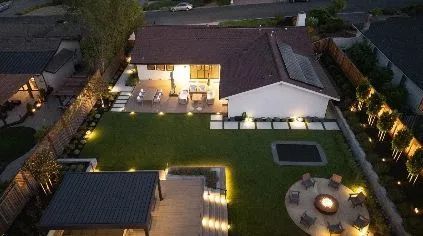 Aerial view of a modern house at dusk with lit patio, landscaped lawn, and circular fire pit seating area