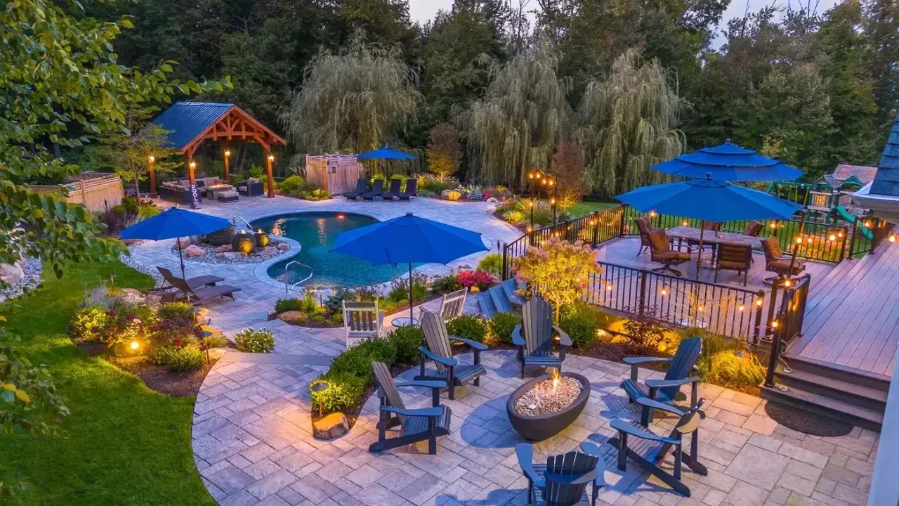 Outdoor resort pool area with blue umbrellas, lounge chairs, fire pit, and lush landscaping at dusk