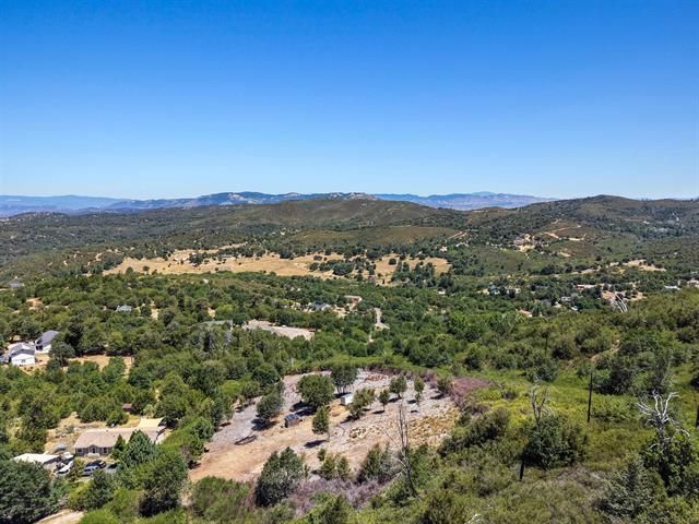 Aerial view of a sprawling, hilly landscape with lush green trees, patches of dry golden terrain, scattered houses, and distant mountain ranges under a clear blue sky.