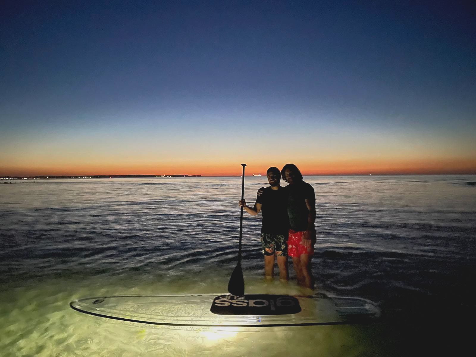 Zwei Personen stehen bei Sonnenuntergang auf einem durchsichtigen Paddelbrett im Meer.