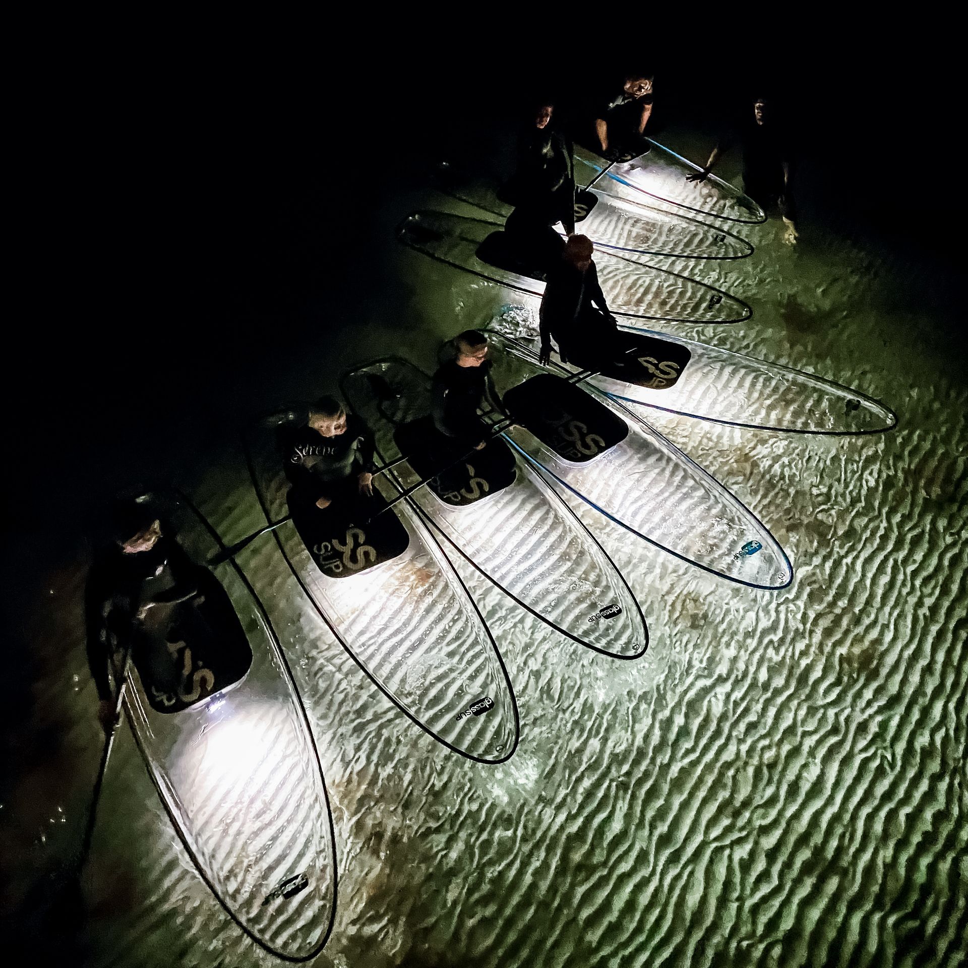 Eine Gruppe von Menschen sitzt nachts auf durchsichtigen Surfbrettern im Wasser