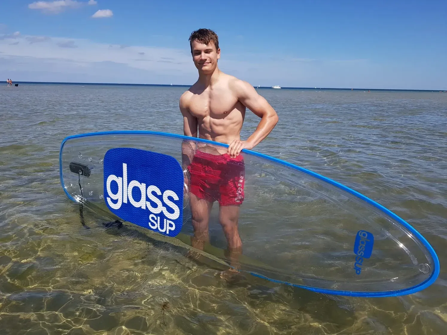Ein Mann ohne Hemd steht im Wasser und hält ein durchsichtiges Surfbrett.
