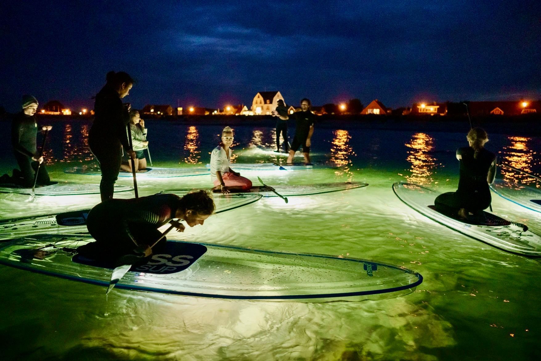 Eine Gruppe von Leuten sitzt nachts auf durchsichtigen Surfbrettern im Wasser.