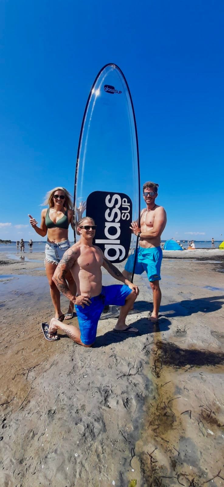 Eine Gruppe von Leuten steht mit einem durchsichtigen Surfbrett in der Hand an einem Strand.