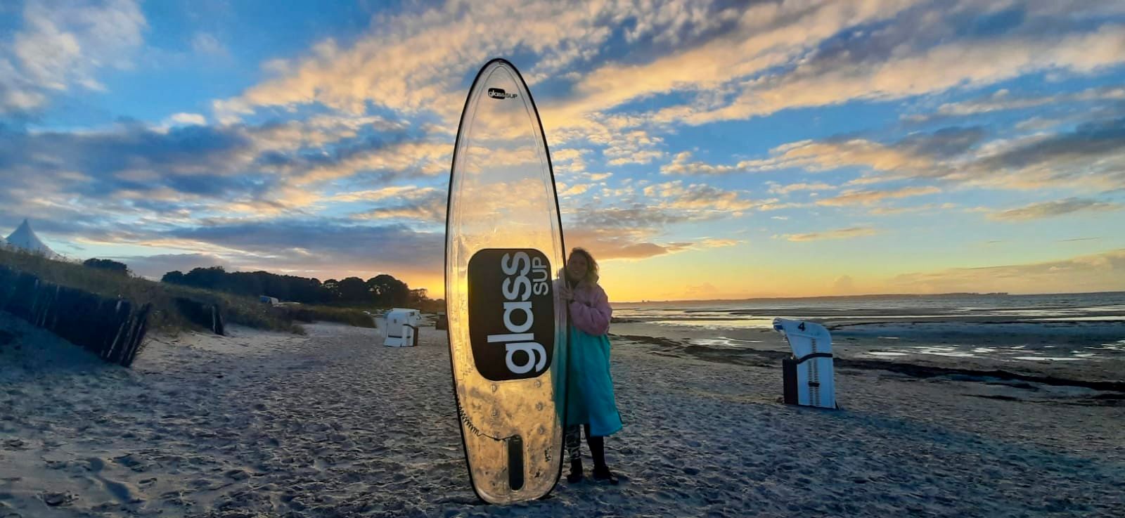 Zwei Personen stehen bei Sonnenuntergang neben einem durchsichtigen Surfbrett an einem Strand.