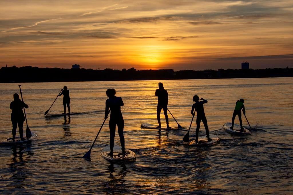 Eine Gruppe von Menschen steht bei Sonnenuntergang auf durchsichtigen Paddelbrettern im Wasser.