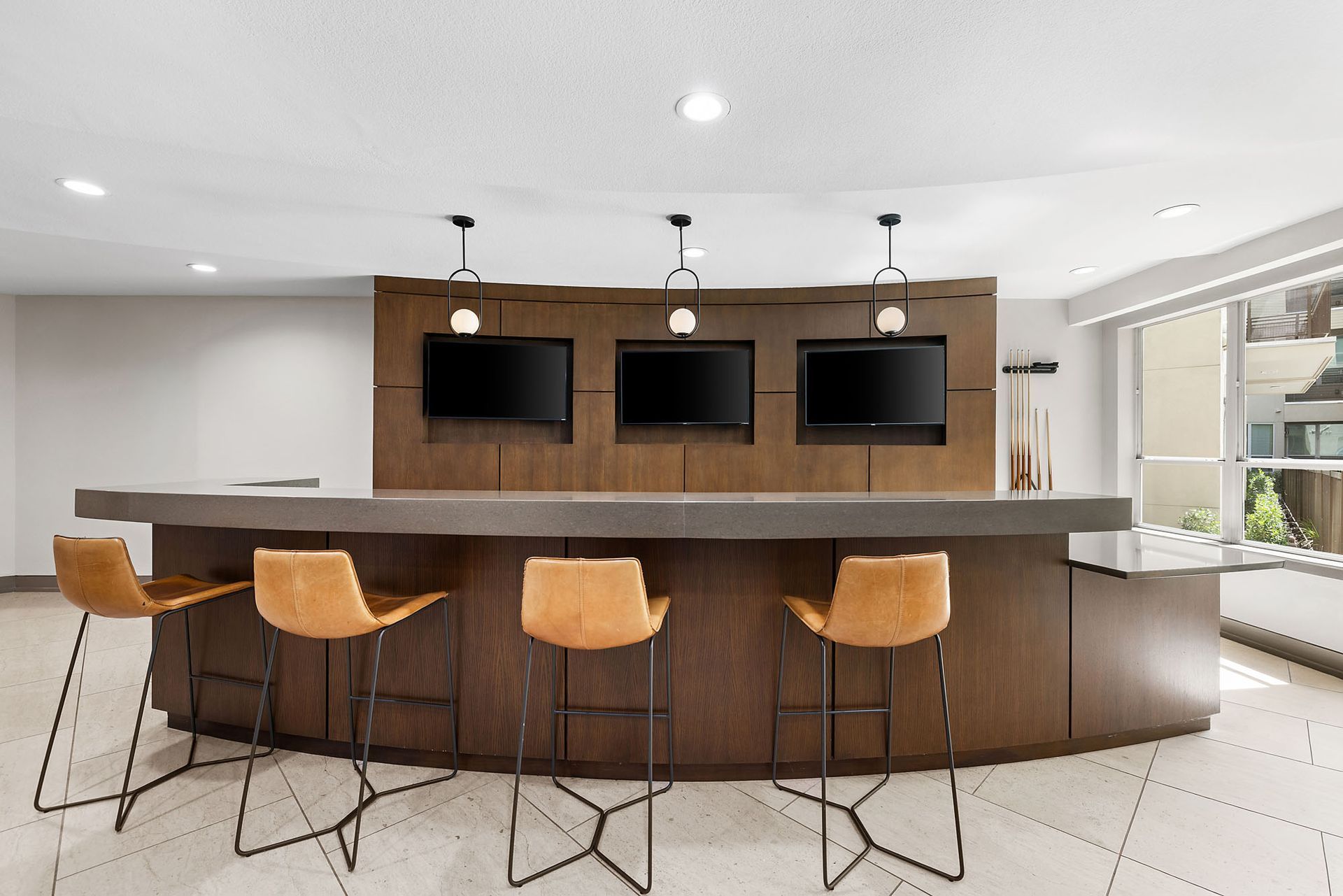 Modern home bar with three wall-mounted TVs, wood-paneled counter, and four stools at Marq on West 7th.