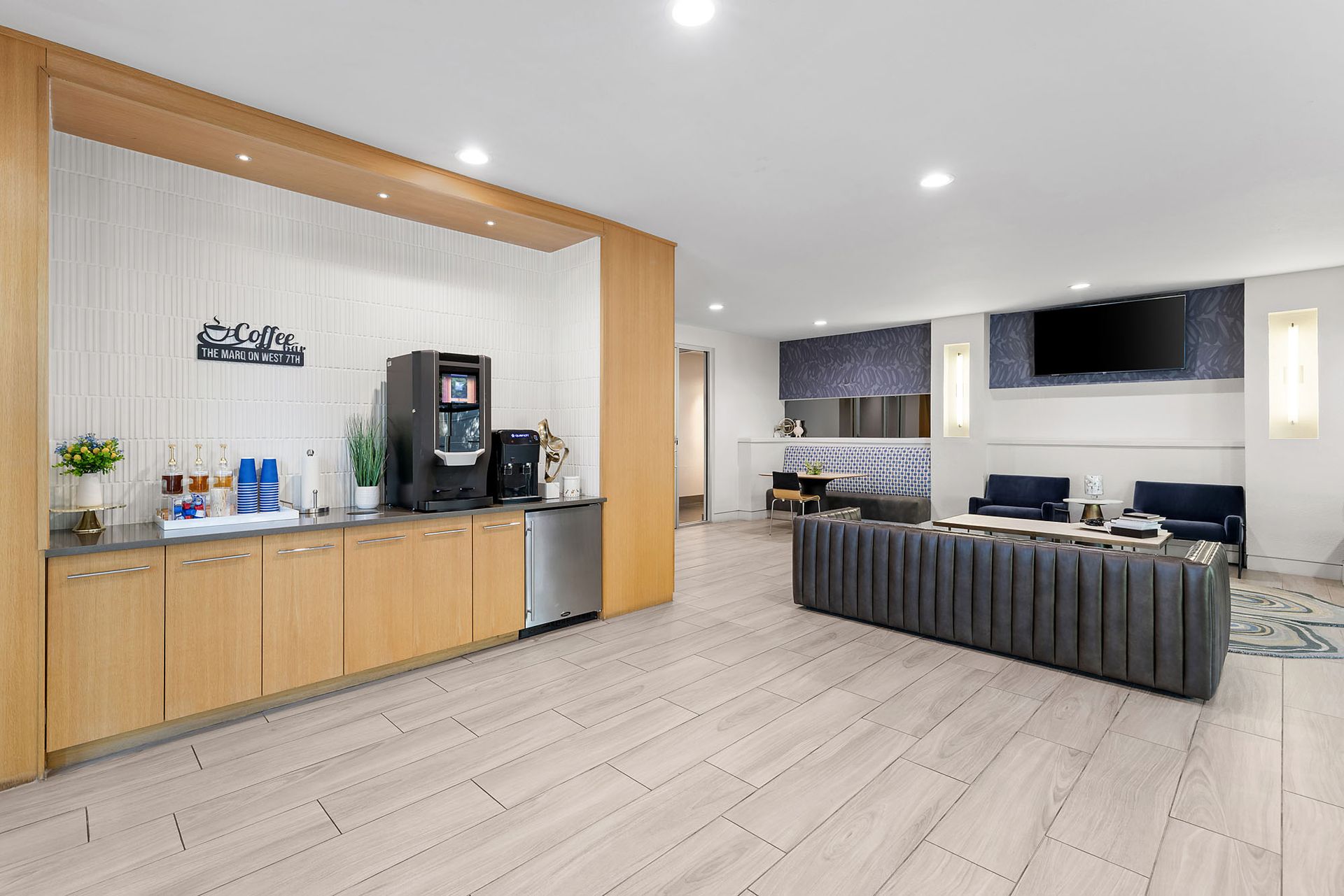 Modern open-plan office lounge with kitchenette, coffee machine, and dark sofas by large windows at Marq on West 7th.