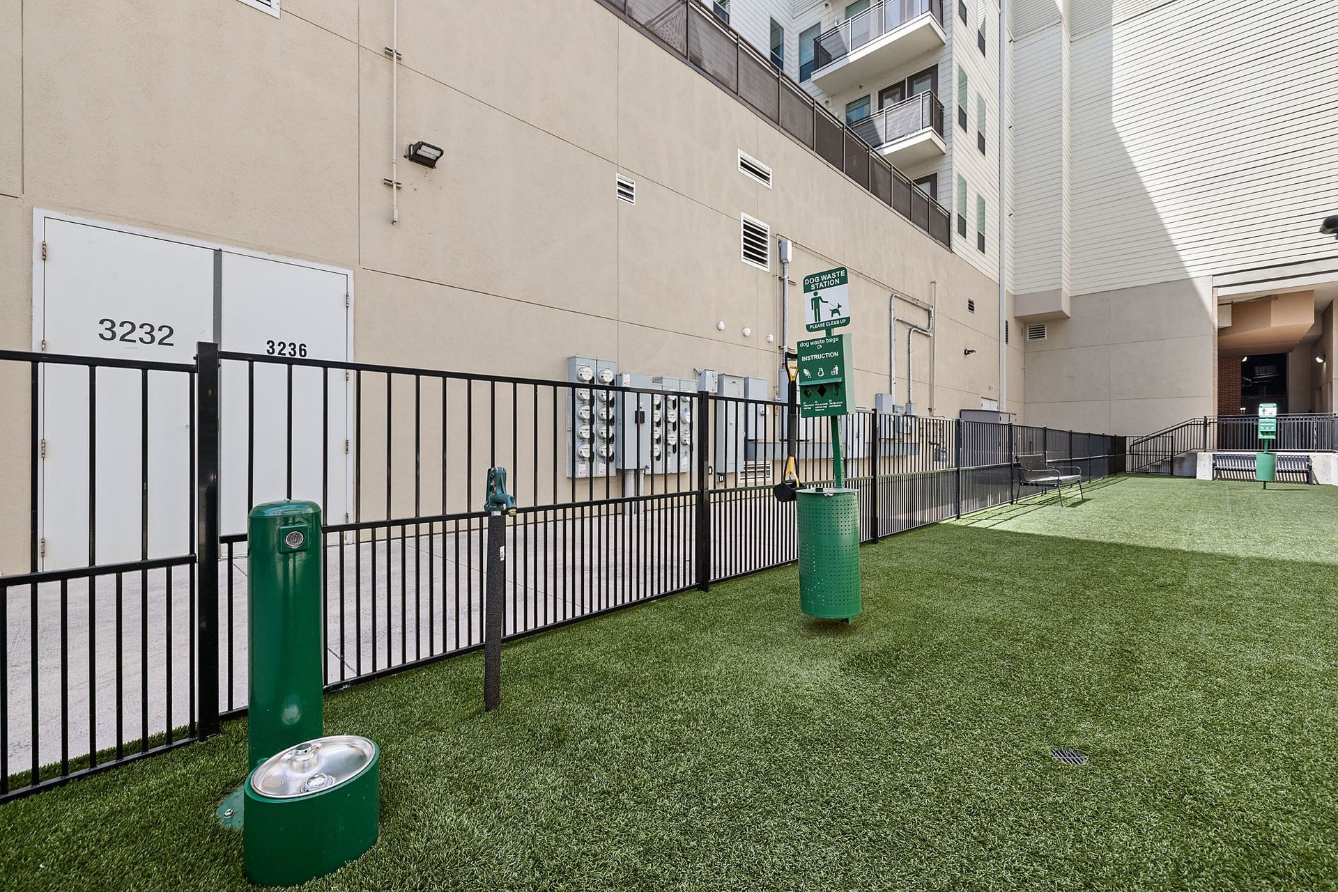 Outdoor courtyard with artificial grass, green posts, and a beige building walkway behind a black fence at Marq on West 7th.