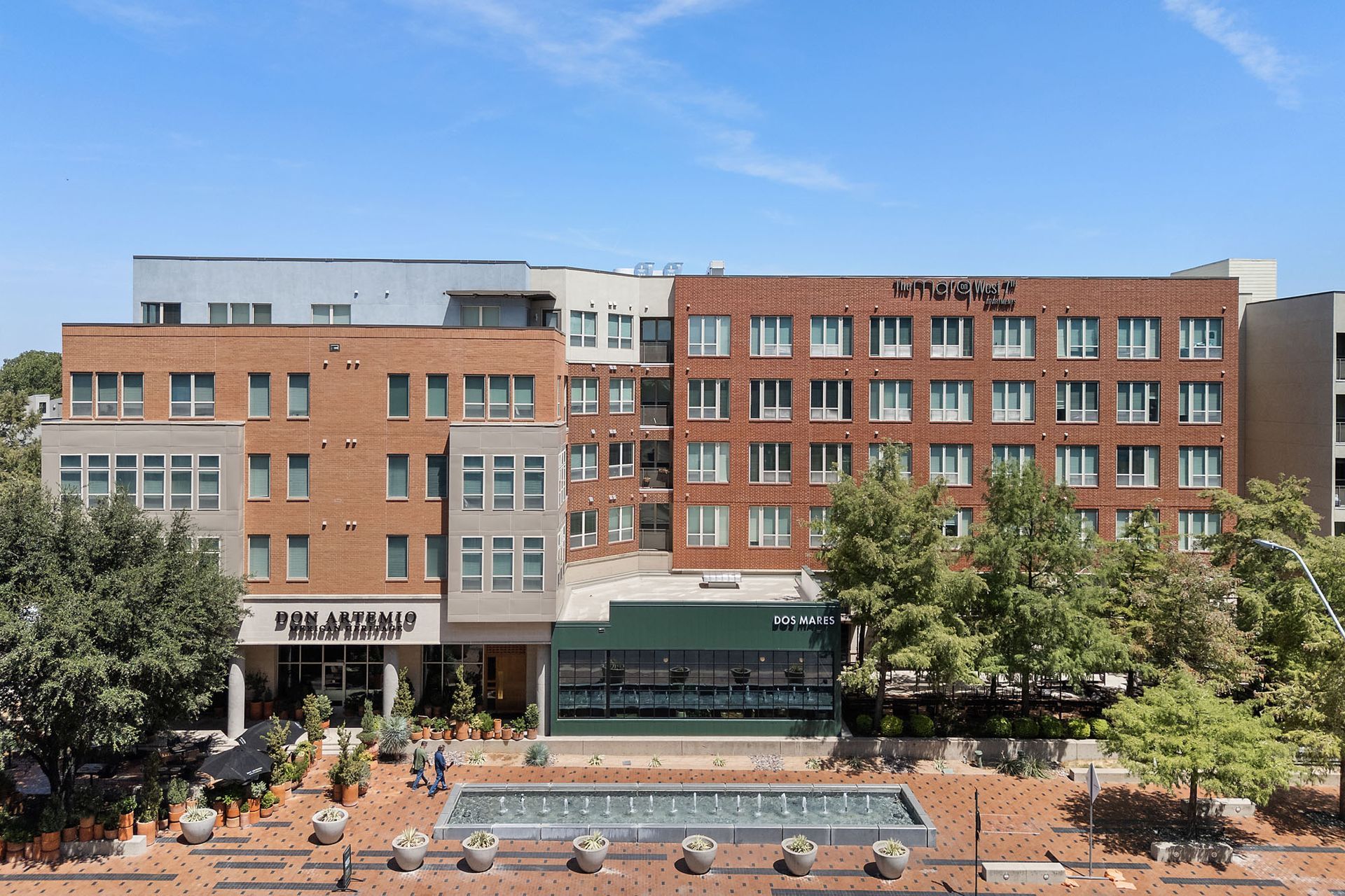 Brick campus building with a central fountain and trees on a sunny day at Marq on West 7th.
