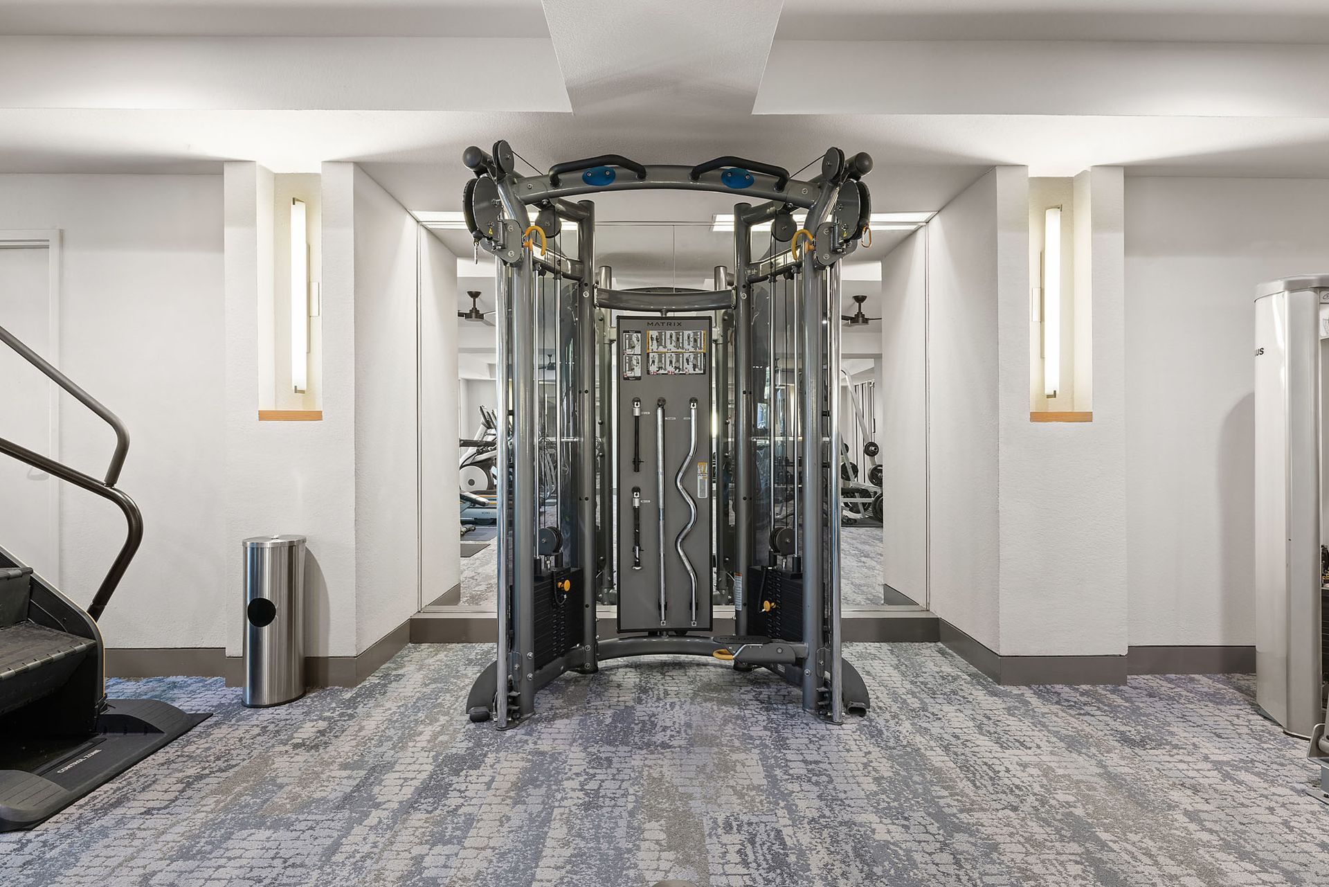 Modern gym with cable workout machine in the center, gray carpet, and white walls in a bright room at Marq on West 7th.
