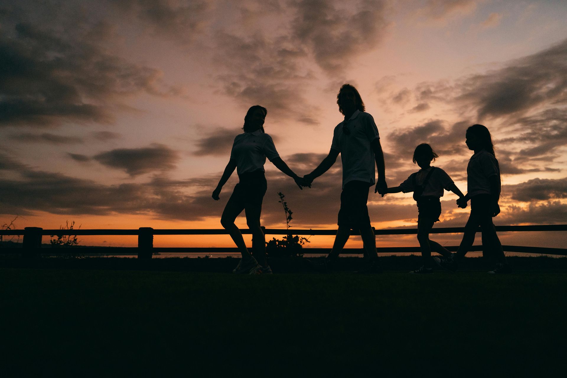 Silhouetted family holding hands on a bridge at sunset under a cloudy sky