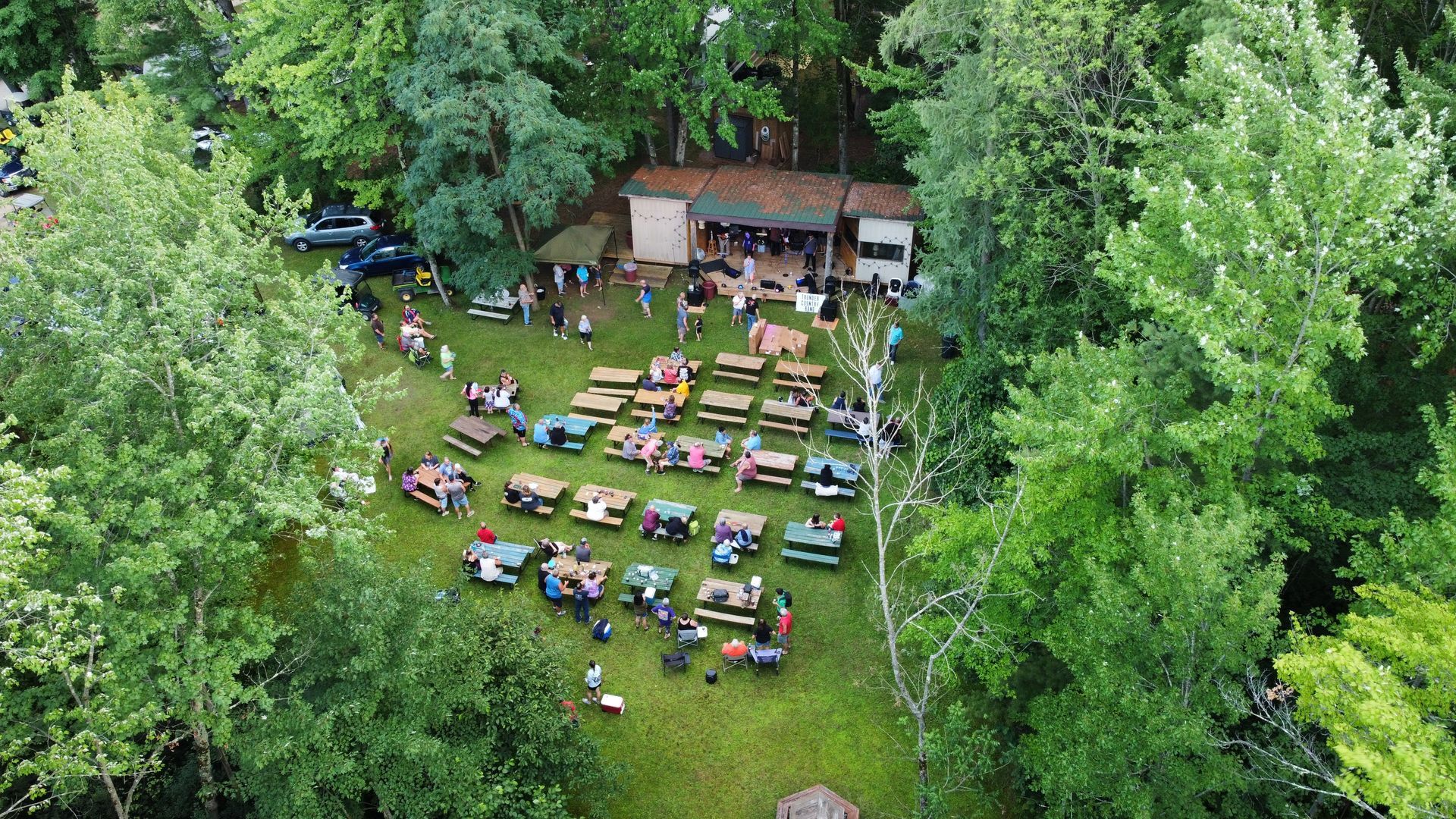 A group of people are sitting at picnic tables in a field surrounded by trees.