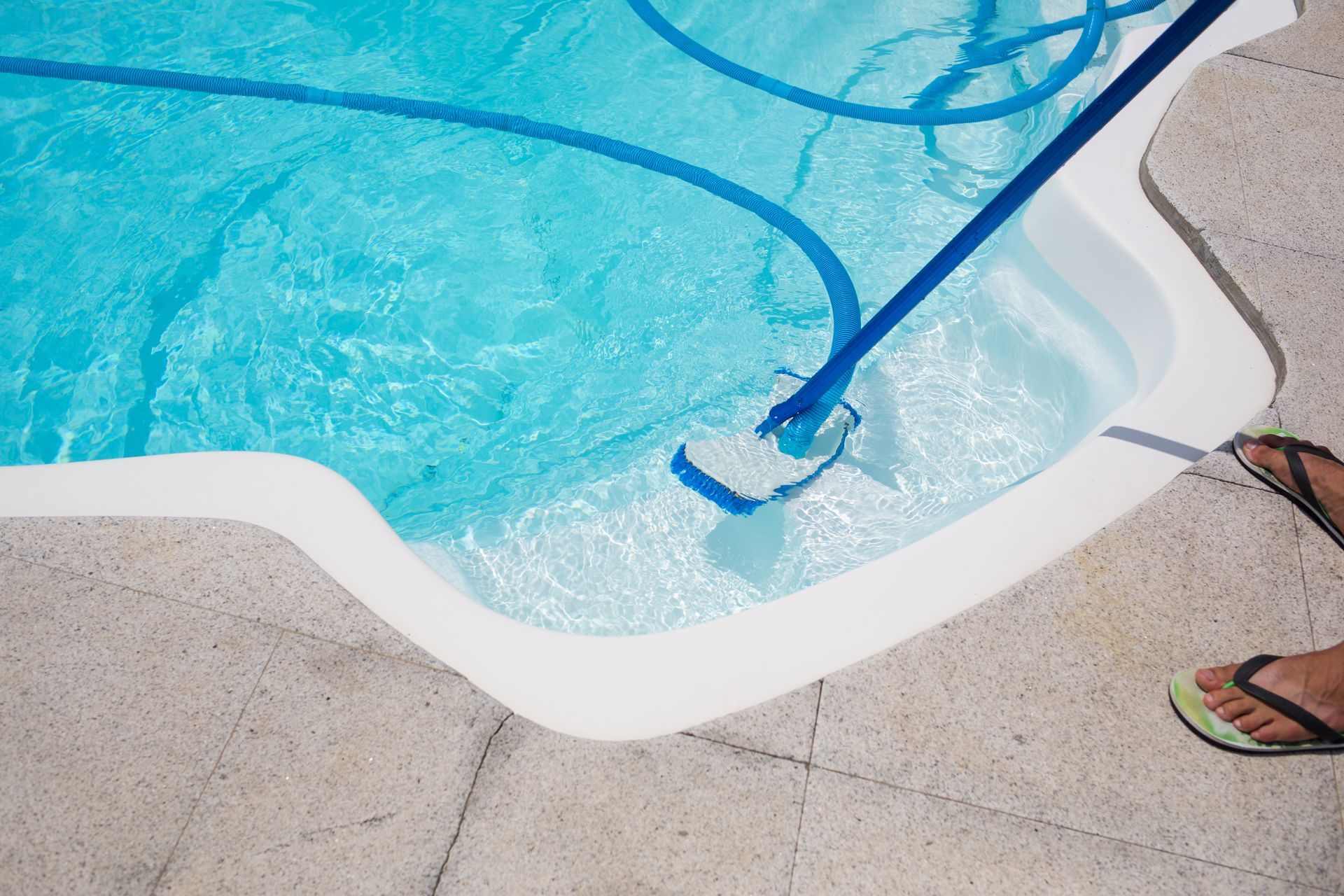 Blue pool vacuum cleaning the edge of a swimming pool, with a foot on the deck.