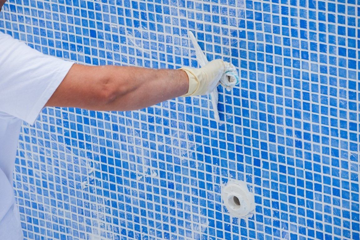 Gloved hand working on a blue mosaic pool wall with a drill or pipe opening
