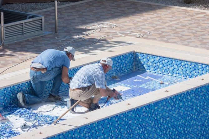 Two workers cleaning a blue-tiled pool with hoses and tools.