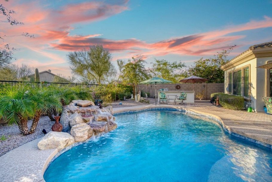 Backyard pool with rock waterfall, landscaped garden, and a pink sunset sky