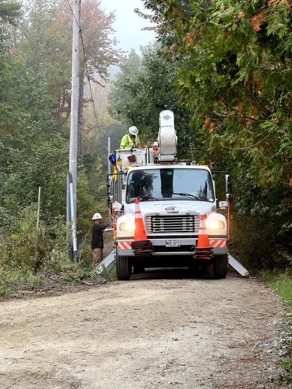 Utility truck with workers on a dirt road in a wooded area, servicing overhead lines.