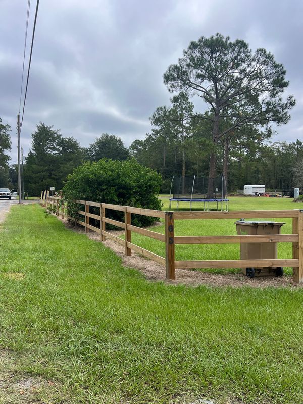Wooden fence along a grassy path under cloudy skies, with trees and a concrete utility box nearby