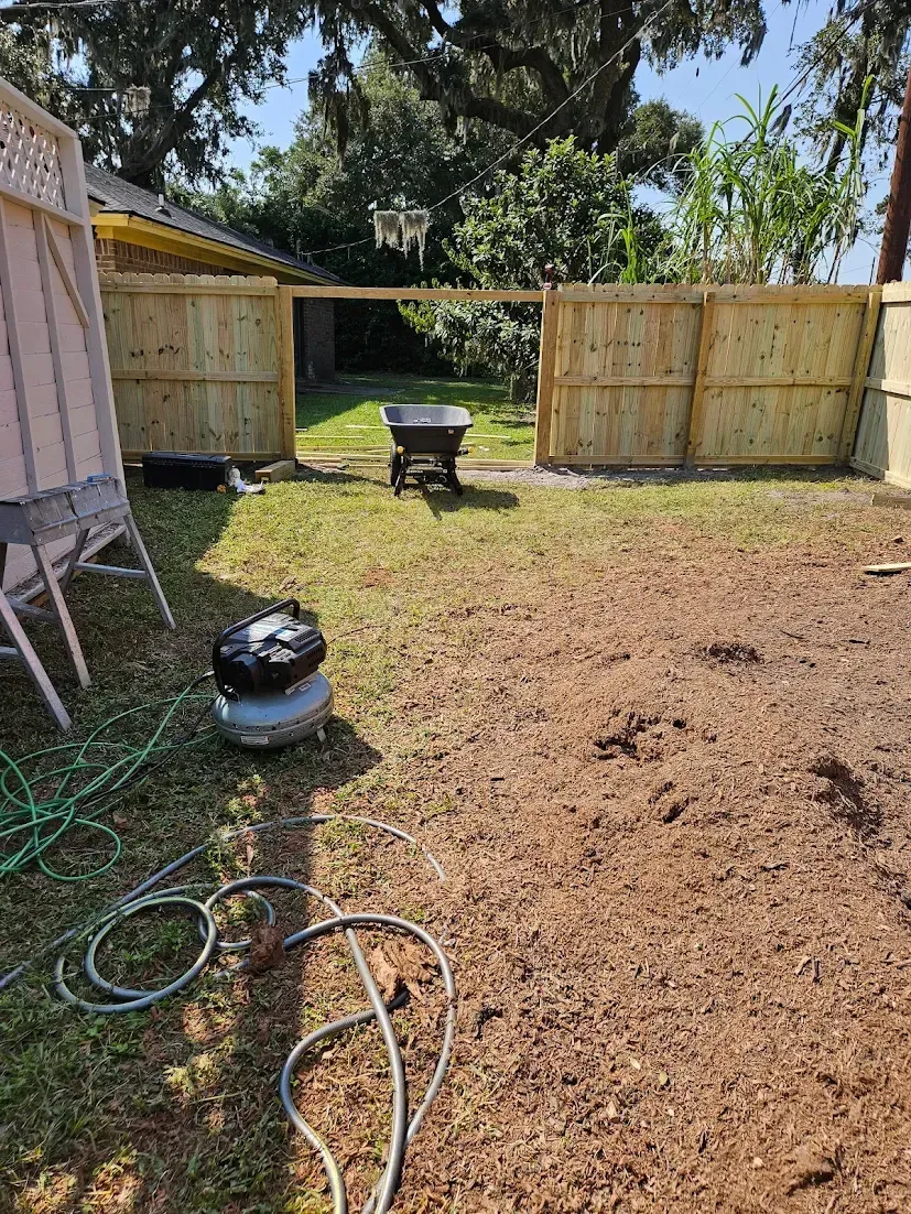 Backyard with fresh dirt and a fenced gate, a wheelbarrow in the center, and tools on the ground in the foreground