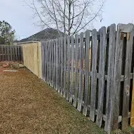 Gray wooden picket fence along a grassy yard with bare trees and a house roof in the background