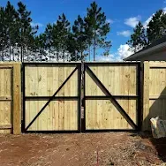 Wooden double gate with black diagonal braces in a fenced yard under blue sky and pine trees
