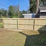 Wooden fence panels along a grassy yard with trees and a house in the background.