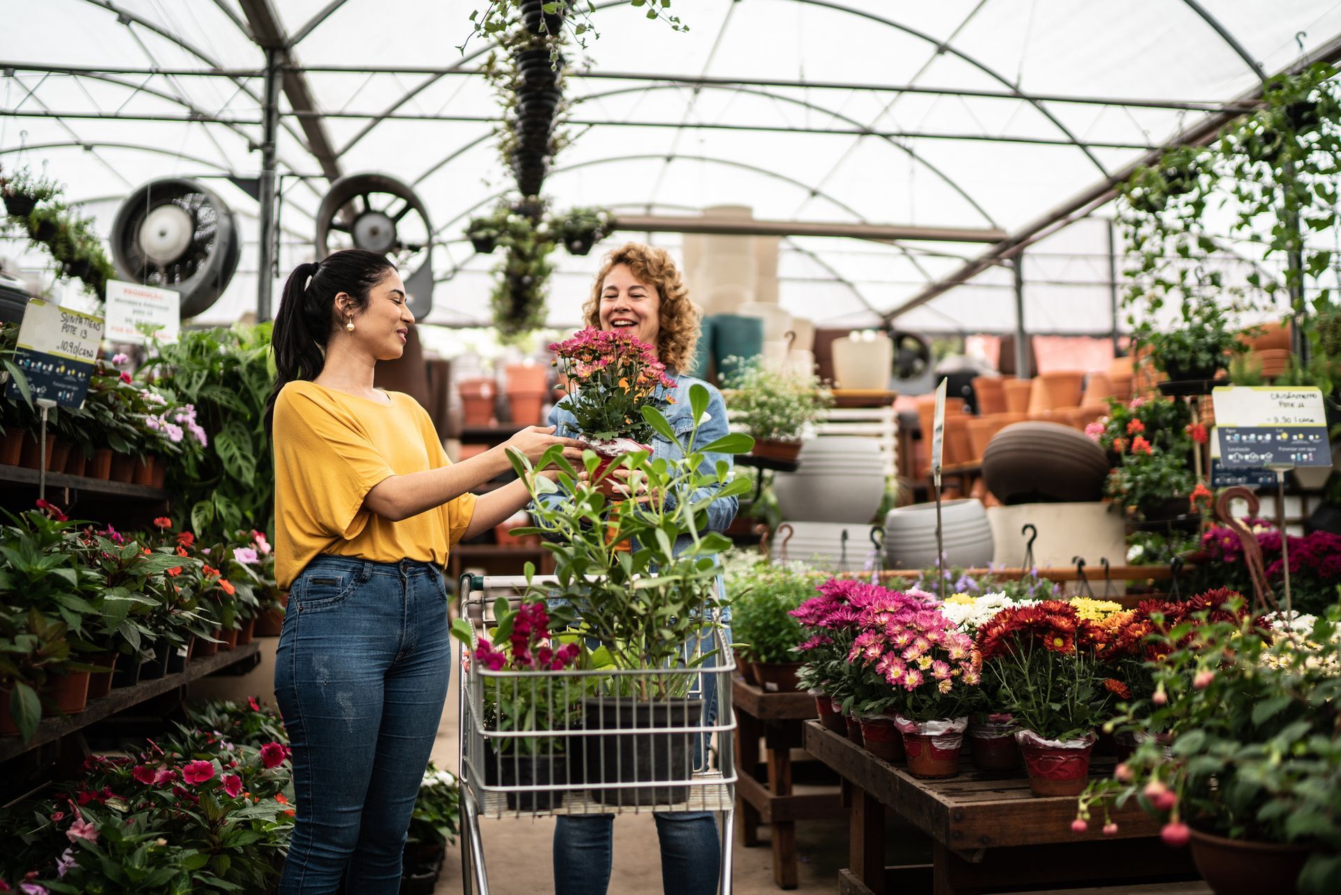 Two women shopping for plants in a greenhouse, adding flowers to a shopping cart.
