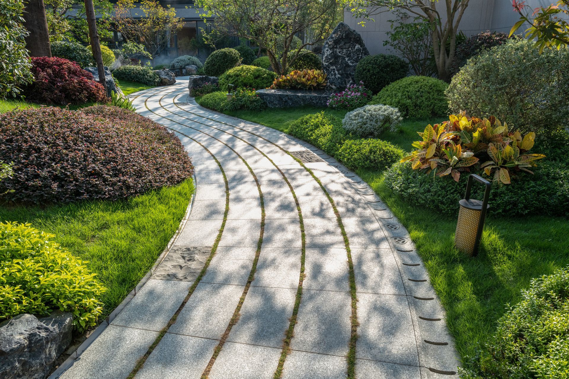 Stone path winding through a lush garden with green grass, colorful bushes, and trees.