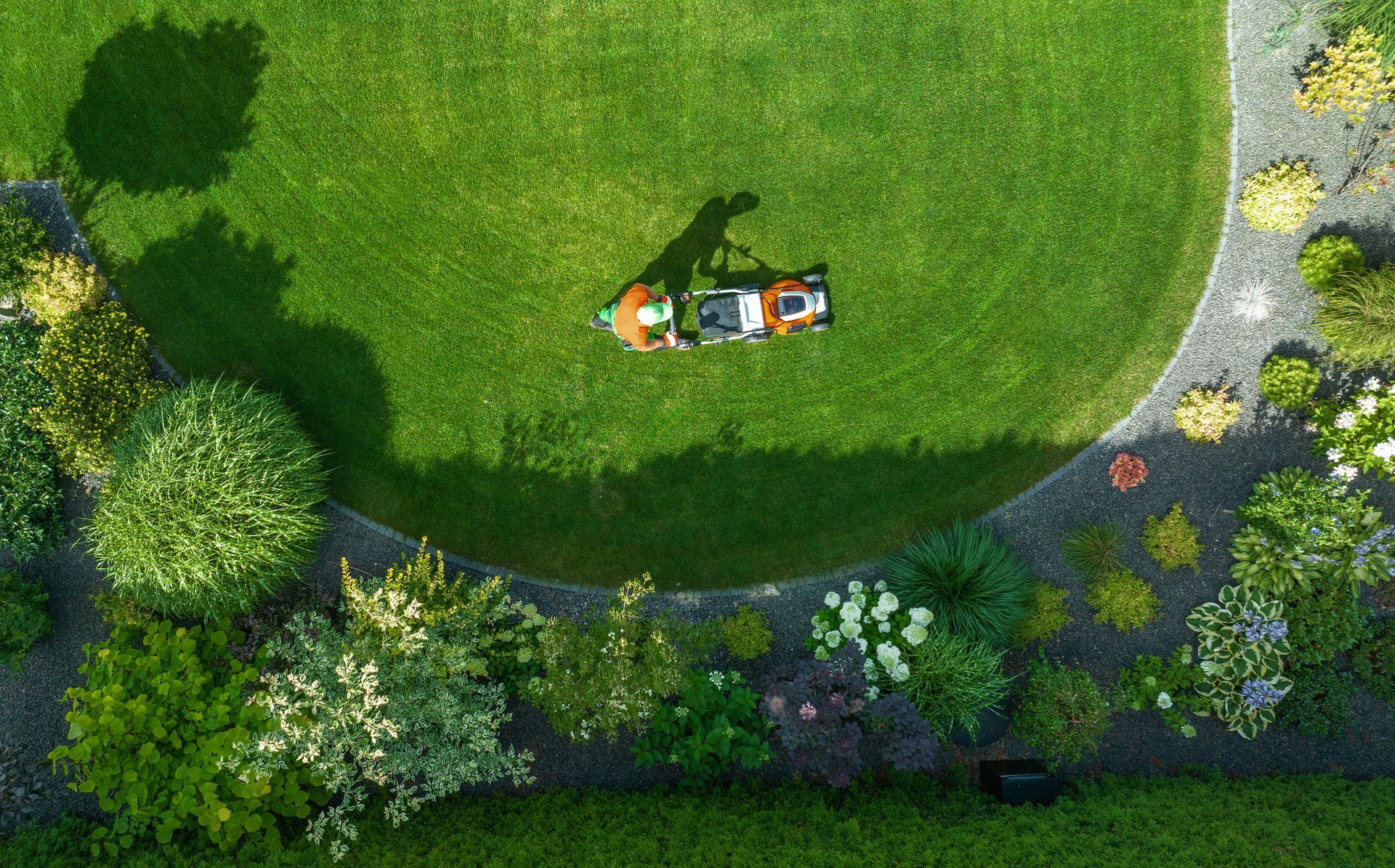 Overhead view of person mowing a green lawn bordered by a stone path and garden with colorful shrubs.