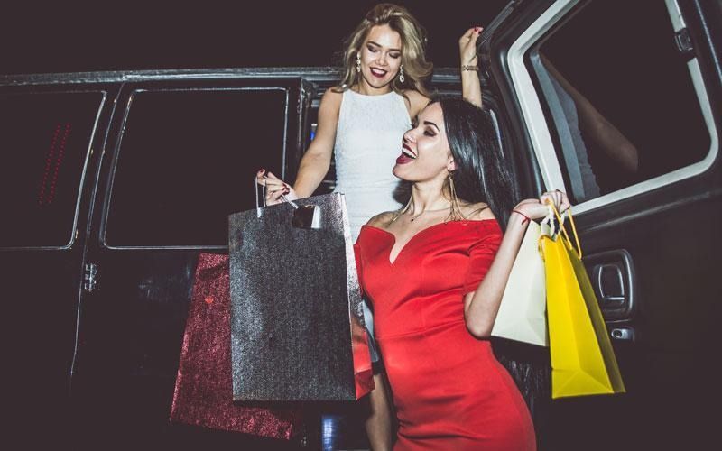 Two women exiting a vehicle with shopping bags, one in a red dress, one in a white dress.