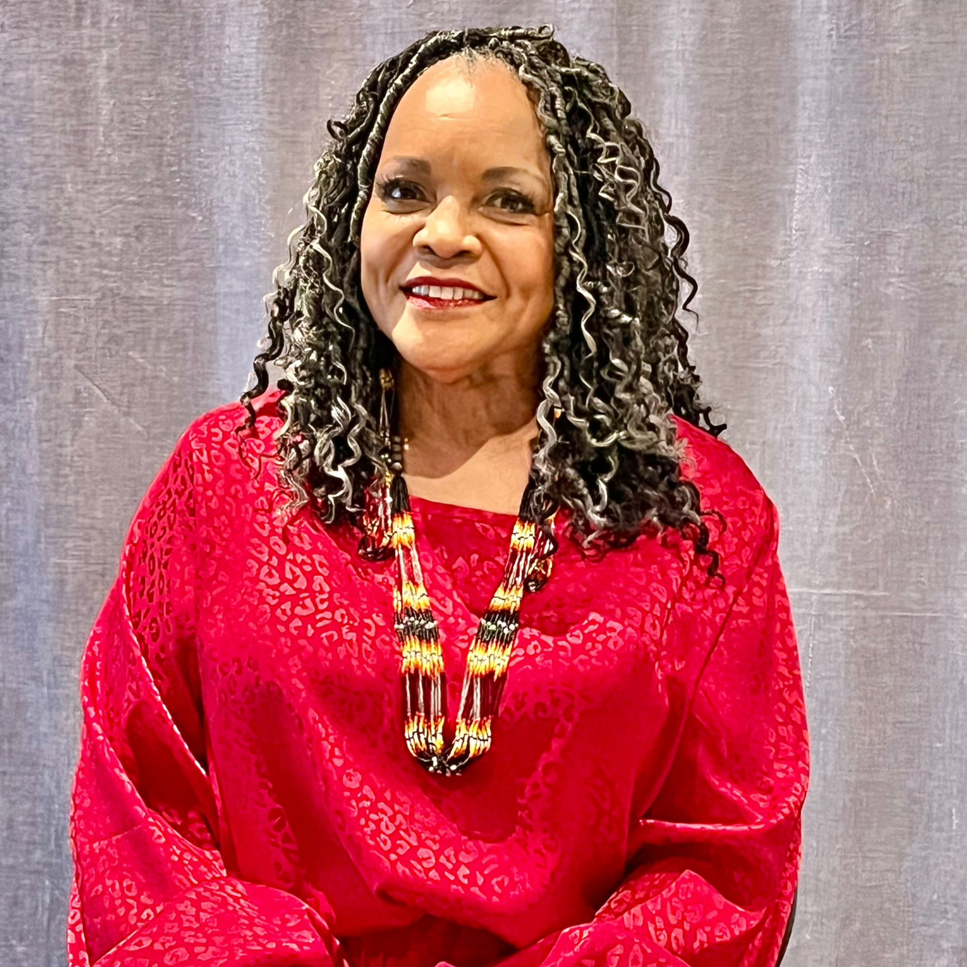 Woman with locs smiles, wearing a red patterned blouse and necklace, standing indoors.