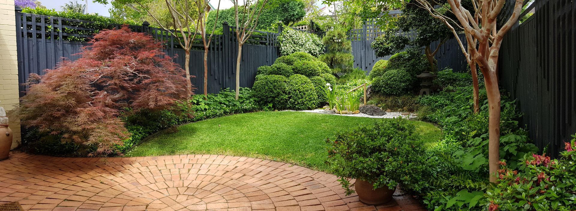 A lush backyard garden with brick patio, manicured green grass, colorful trees, and a dark wooden fence.