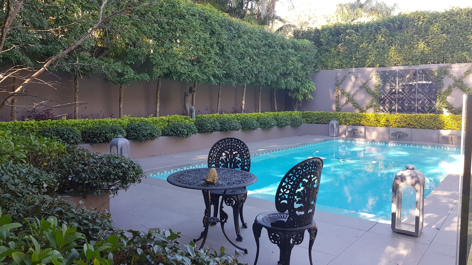 Poolside scene with wrought-iron table and chairs, lush greenery, and a water feature.