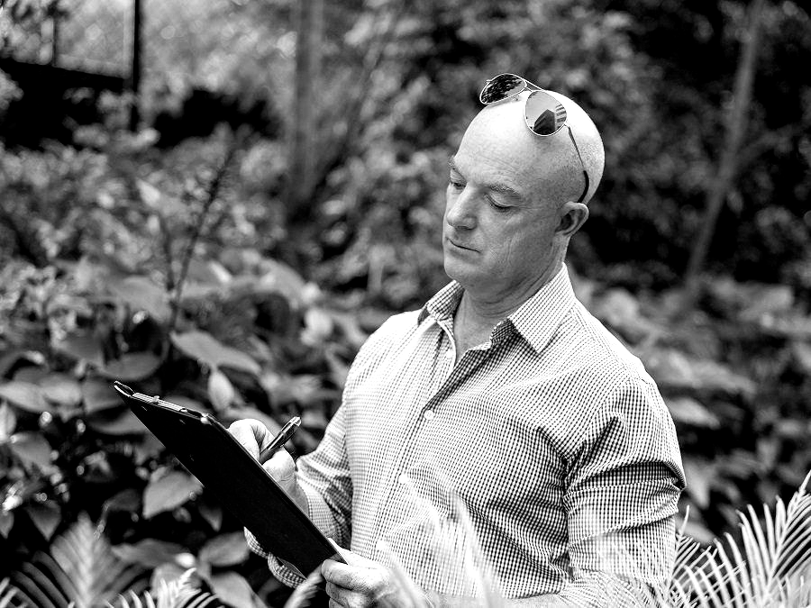 Bald man wearing sunglasses on head, writing on a clipboard outdoors amongst plants.