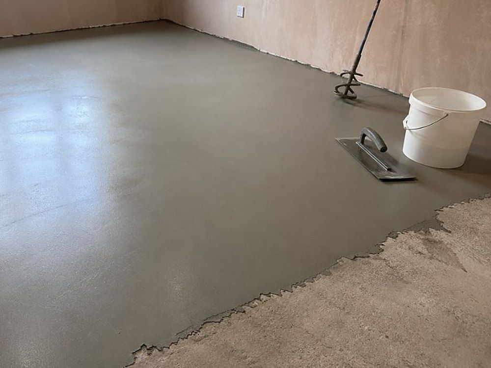 Freshly poured levelling compound across a floor, with a trowel, mixing paddle and bucket beside it