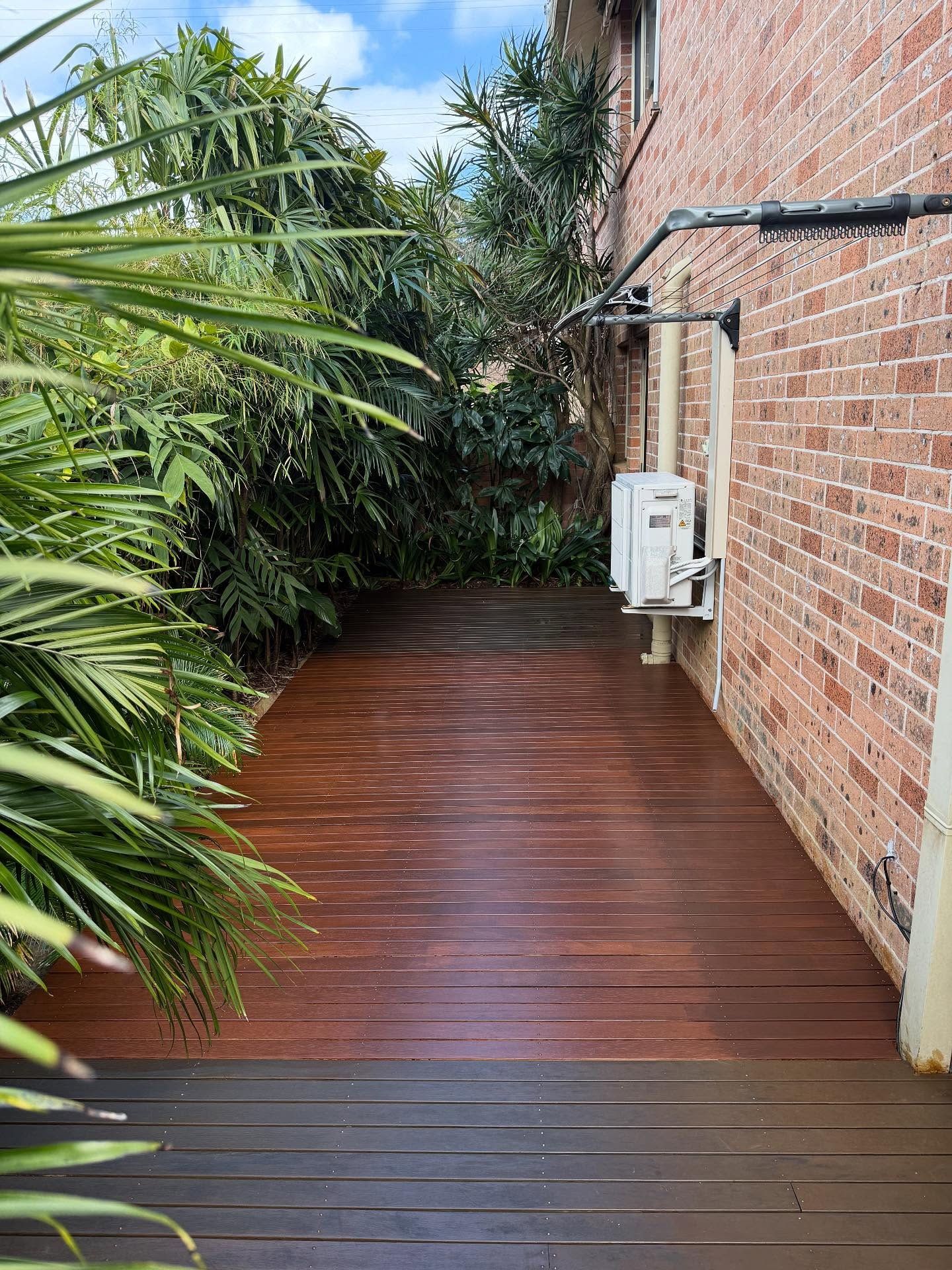 Wooden walkway beside a brick house, bordered by lush tropical greenery and a small outdoor unit — Tyrone Building Solutions in Salamander Bay, NSW