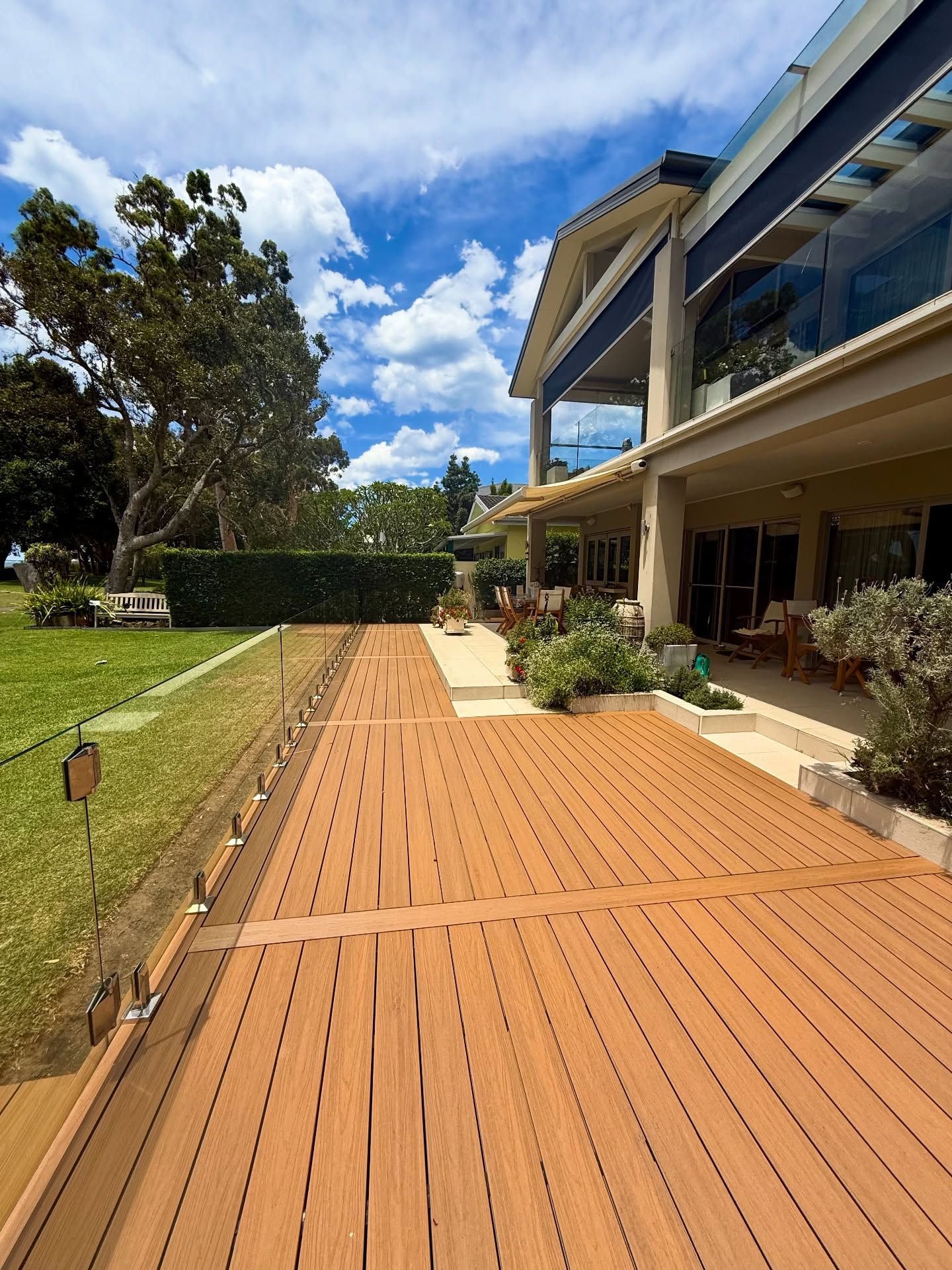 Wooden deck path beside a house with potted plants, green lawn, hedges, and a bright blue sky — Tyrone Building Solutions in Anna Bay, NSW