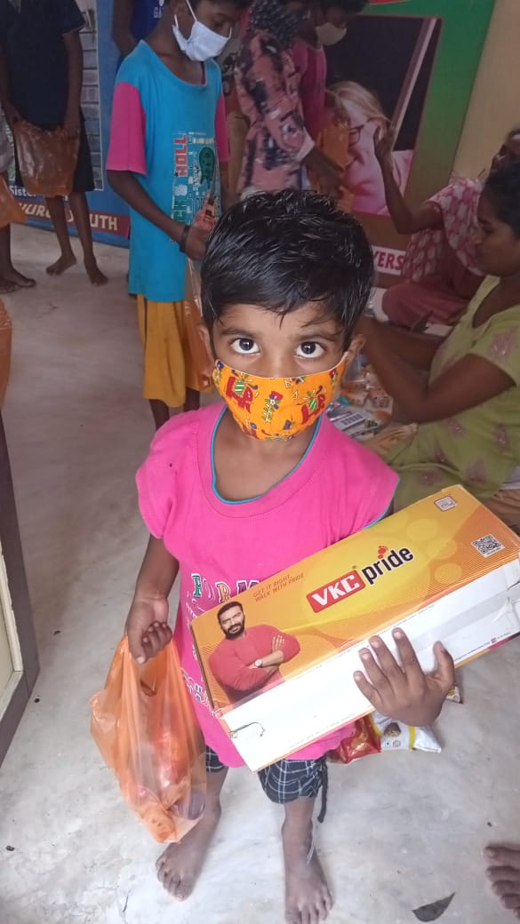 Kind met masker houdt schoenen en tas vast; mensen op de achtergrond. Binnen, felle kleuren.